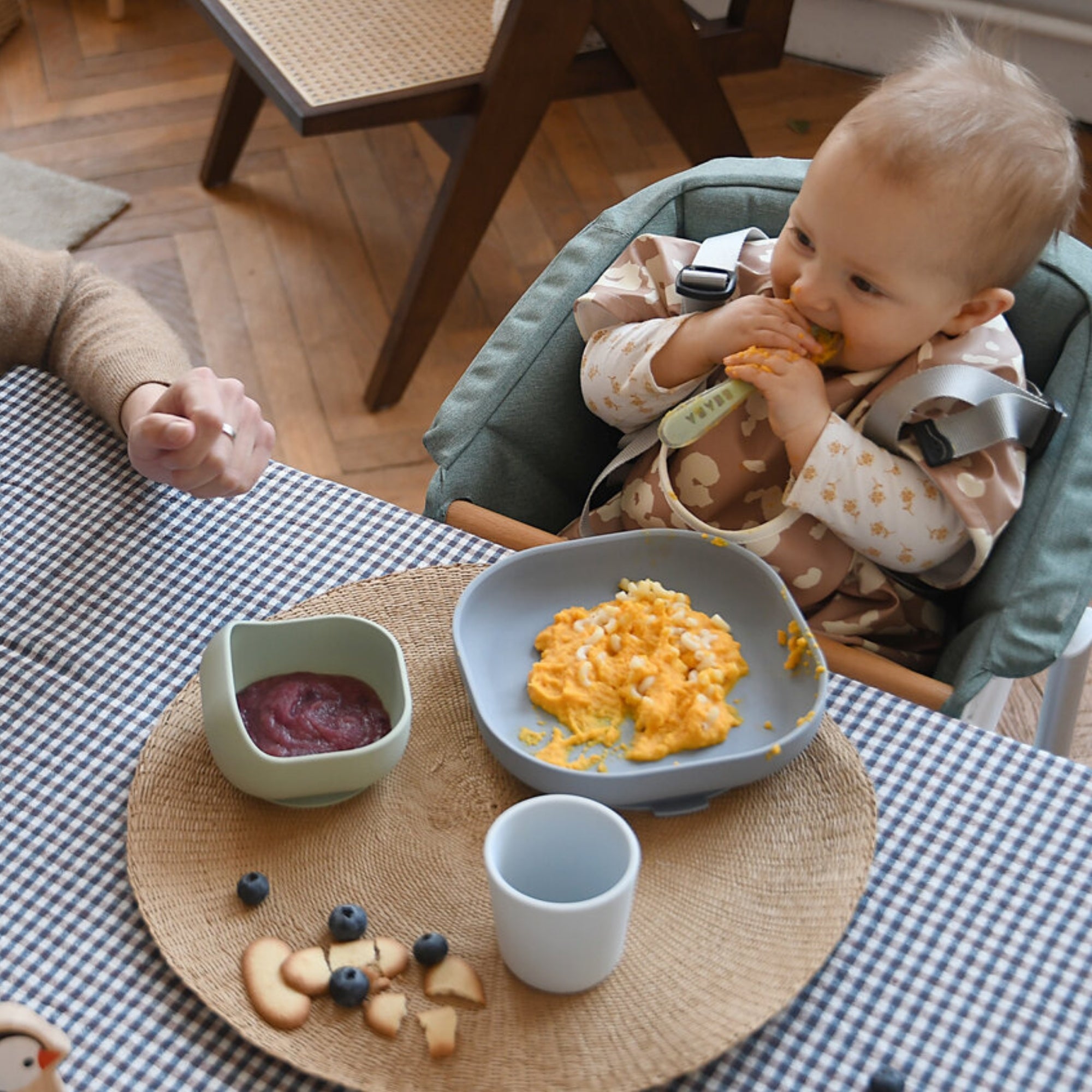 baby in highchair holding a spoon to his mouth, with a Beaba silicone suction meal set filled with food in front of him